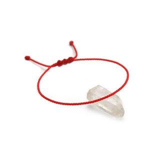 Peut inclure: Un bracelet en cordon rouge avec une fermeture à nœud réglable. Un cristal clair et pointu est attaché au bracelet. Le bracelet est présenté sur un fond blanc.