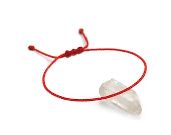 Könnte beinhalten: Ein rotes Schnurarmband mit verstellbarem Knotenverschluss. Ein klarer, spitzer Kristall ist an dem Armband befestigt. Das Armband wird auf weißem Hintergrund präsentiert.