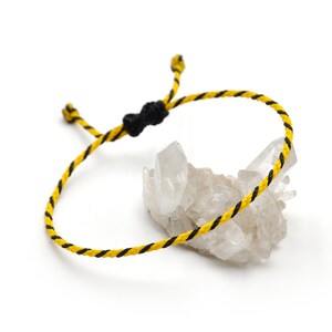 Könnte beinhalten: Ein geflochtenes Armband in Gelb und Schwarz mit einem schwarzen Knotenverschluss. Das Armband liegt auf einer weißen Oberfläche mit einem klaren Quarzkristall-Cluster.