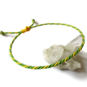 May include: A green and yellow braided cord bracelet with a sliding knot fastening. The bracelet is resting on a cluster of clear quartz crystals.
