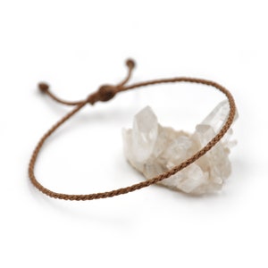 May include: A brown braided cord bracelet with an adjustable closure. The bracelet is lying on a white surface with a cluster of clear quartz crystals.