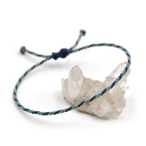 Peut inclure: Un bracelet tressé avec des fils bleus, turquoise et gris. Le bracelet est noué et repose sur un groupe de cristaux de quartz transparents.