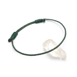 Op de afbeelding: Een donkergroene verstelbare armband met een schuifknoopsluiting. De armband is gemaakt van een dun, geweven koord. Een heldere bergkristal ligt naast de armband op een witte achtergrond.