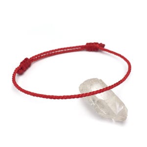 May include: A red string bracelet with adjustable knots. A clear quartz crystal is next to the bracelet. The bracelet is a simple design, likely for everyday wear. The background is white.