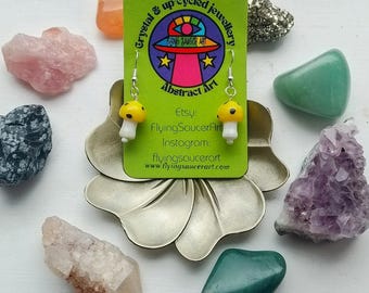 May include: A pair of mushroom-shaped earrings with yellow caps and white stems, hanging from silver hooks. The earrings are displayed on a green card with the text "Crystal & up-cycled jewellery" and "Flying Saucer Art". Various crystals and gemstones surround the earrings.