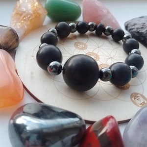 May include: A black beaded bracelet featuring large, round beads and smaller, silver-coloured beads. The bracelet is displayed on a wooden coaster with a floral design, surrounded by colourful gemstones.