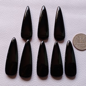May include: Eight polished, black teardrop-shaped gemstones are arranged on a light surface. A U.S. quarter coin is included for size comparison. The gemstones have a glossy finish and a smooth texture. These could be used for jewelry making or other crafts.