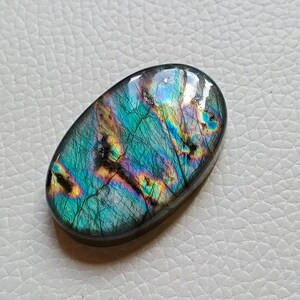 May include: An oval-shaped labradorite gemstone featuring a polished, smooth surface with iridescent flashes of blue, green, and gold. The stone displays natural internal fractures and dark inclusions that contrast with the vibrant, reflective color play.