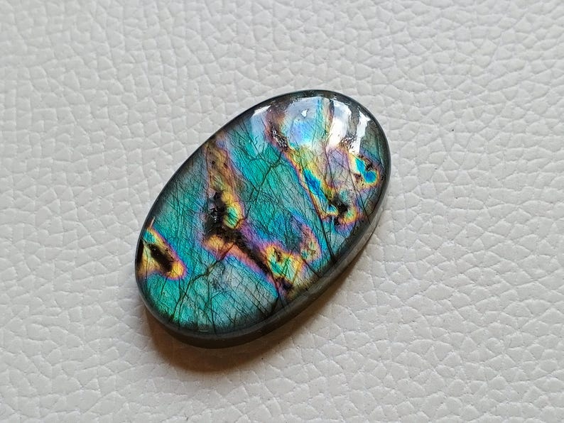 May include: An oval-shaped labradorite gemstone featuring a polished, smooth surface with iridescent flashes of blue, green, and gold. The stone displays natural internal fractures and dark inclusions that contrast with the vibrant, reflective color play.