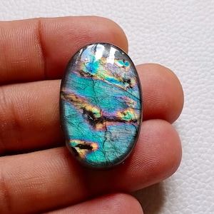 May include: An oval-shaped labradorite gemstone featuring a polished surface with iridescent flashes of teal, blue, and gold. The stone displays natural internal fractures and dark inclusions that contrast with the vibrant, multi-colored sheen.
