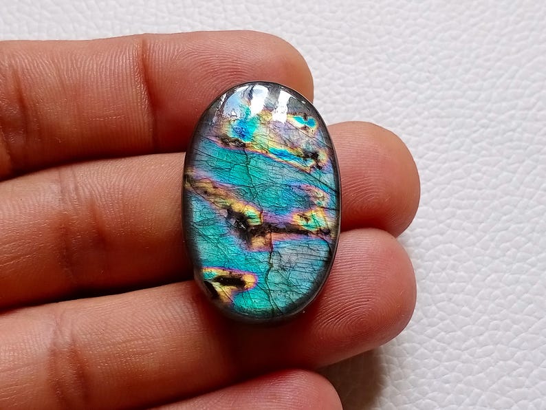 May include: An oval-shaped labradorite gemstone featuring a polished surface with iridescent flashes of teal, blue, and gold. The stone displays natural internal fractures and dark inclusions that contrast with the vibrant, multi-colored sheen.
