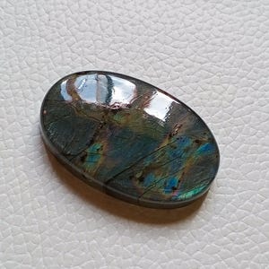 May include: An oval-shaped labradorite gemstone featuring a polished, smooth surface with iridescent flashes of blue, green, and gold colors. The stone displays natural internal fractures and dark grey tones throughout its crystalline structure.