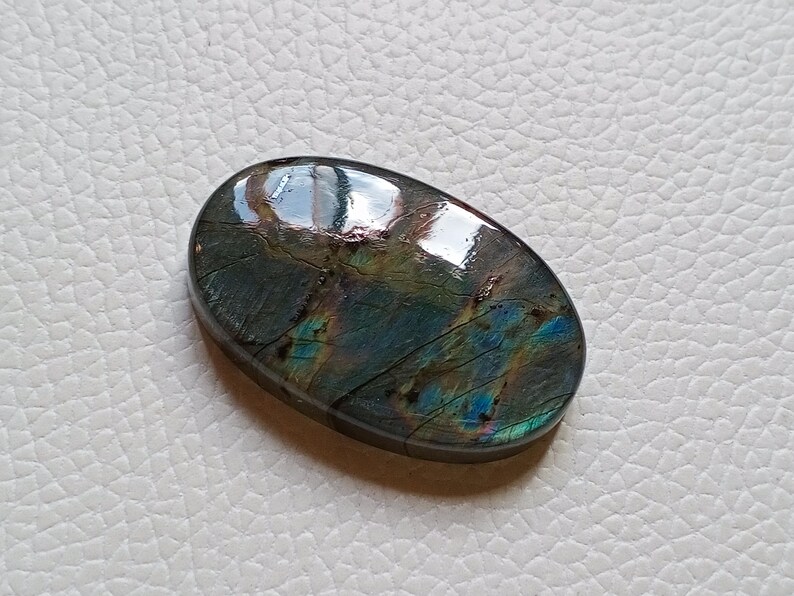 May include: An oval-shaped labradorite gemstone featuring a polished, smooth surface with iridescent flashes of blue, green, and gold colors. The stone displays natural internal fractures and dark grey tones throughout its crystalline structure.