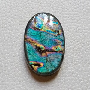 May include: An oval-shaped labradorite gemstone featuring a polished surface with iridescent flashes of teal, blue, and gold. The stone displays natural internal fractures and dark inclusions throughout its structure, creating a vibrant play of color.