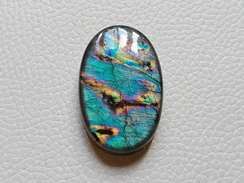 May include: An oval-shaped labradorite gemstone featuring a polished surface with iridescent flashes of teal, blue, and gold. The stone displays natural internal fractures and dark inclusions throughout its structure, creating a vibrant play of color.