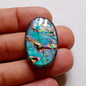May include: An oval-shaped labradorite gemstone featuring a polished surface with iridescent flashes of teal, blue, and gold. The stone displays natural internal fractures and a smooth, rounded edge, showcasing a vibrant play of color.
