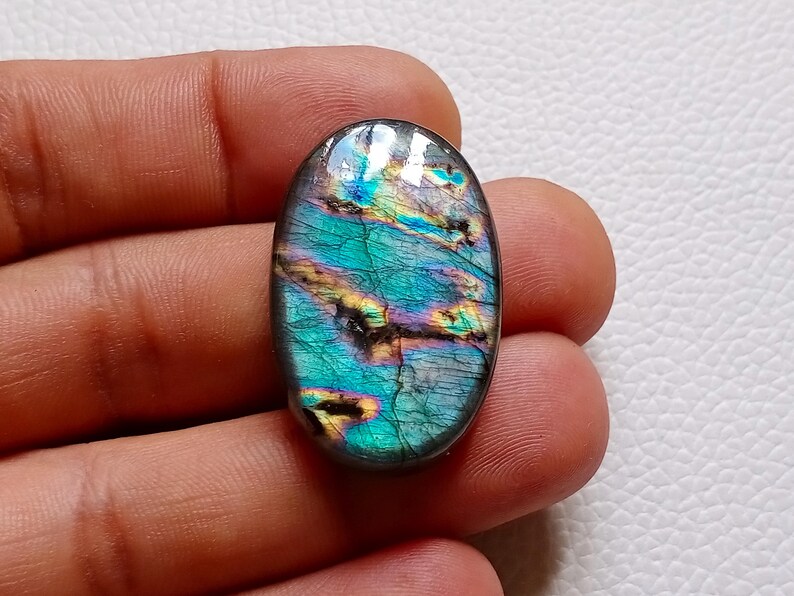 May include: An oval-shaped labradorite gemstone featuring a polished surface with iridescent flashes of teal, blue, and gold. The stone displays natural internal fractures and a smooth, rounded edge, showcasing a vibrant play of color.