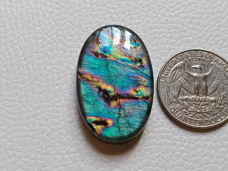 May include: An oval-shaped labradorite gemstone featuring iridescent flashes of blue, green, and yellow with dark internal inclusions. The stone is positioned next to a United States quarter coin for scale, showing its relative size.