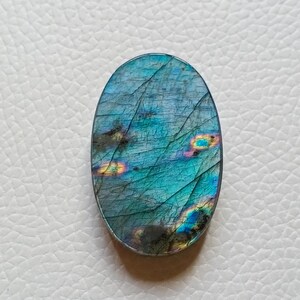 May include: An oval-shaped labradorite gemstone featuring a polished surface with a deep blue and teal base color. The stone displays iridescent flashes of yellow, orange, and green light across its textured, cracked, and layered crystalline structure.