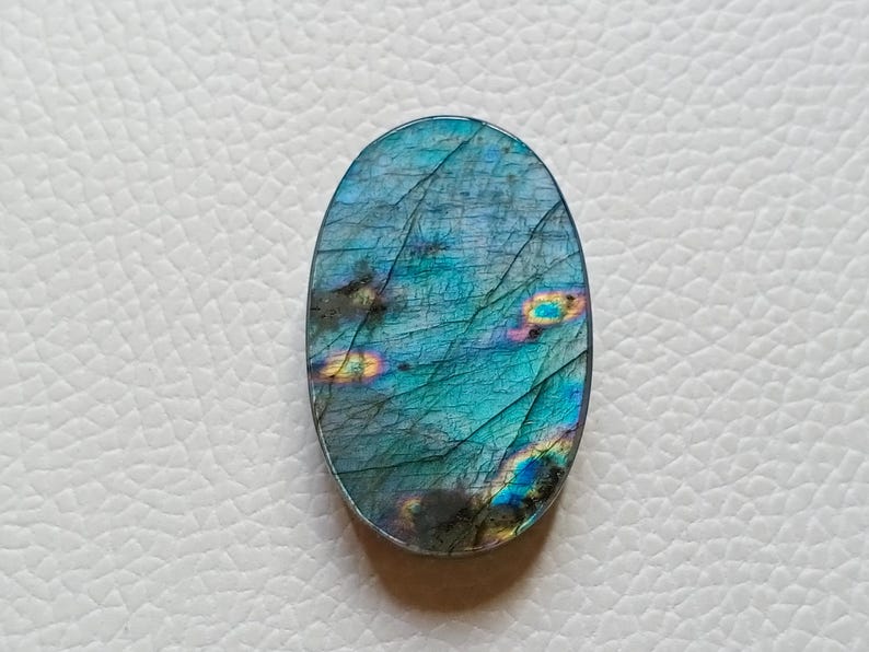 May include: An oval-shaped labradorite gemstone featuring a polished surface with a deep blue and teal base color. The stone displays iridescent flashes of yellow, orange, and green light across its textured, cracked, and layered crystalline structure.