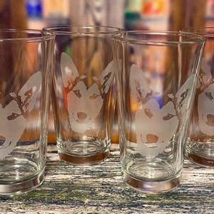 May include: Four clear glass tumblers with a frosted wolf head design. The glasses are cylindrical with a slightly flared base. The wolf design is etched into the glass, creating a contrasting white image against the clear glass.