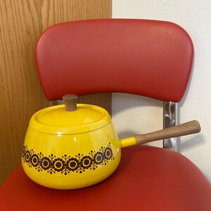 May include: A yellow saucepan with a wooden handle and lid knob, decorated with a brown floral pattern. The saucepan is set against a red chair.