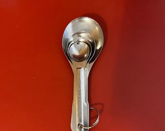 May include: A set of three nesting stainless steel measuring spoons with a ring handle. The spoons are silver and have a shiny finish.