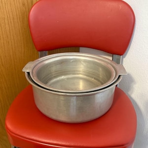 May include: A vintage aluminum cooking pot with a lid sits on a red vinyl chair. The chair has a rounded back and chrome legs. The pot is round with two handles and a silver finish. The background includes a wooden door and a white wall.