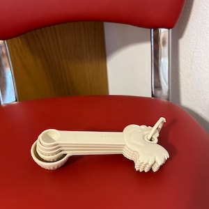 May include: A set of off-white measuring spoons with a decorative elephant-shaped handle. The spoons are stacked and rest on a red surface. Each spoon is labeled with a measurement, such as 1/4 tsp.