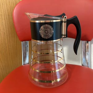 May include: Vintage glass pitcher with a black top and handle, accented with gold trim and horizontal stripes. The black top features a gold floral design. The pitcher is likely used for serving beverages.