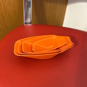 May include: A stack of orange plastic measuring scoops. The scoops are nested together, showing different sizes. The scoops are on a red surface, with a red chair and a wooden wall in the background.