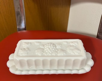 Puede incluir: Mantequillera de vidrio blanco con tapa rectangular con diseño de uvas y hojas. La base tiene un borde festoneado. El plato está sobre una superficie roja.