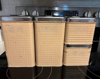 May include: A set of vintage kitchen canisters in a light tan color with a checkered pattern. The canisters are labeled "Sugar", "Flour", "Tea", and "Coffee" in embossed lettering. Each canister has a silver-colored metal lid.