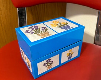 Può includere: Una scatola per ricette in metallo blu con un design floreale. La scatola ha tre scomparti ed è decorata con immagini ricamate di fiori in vaso.