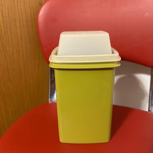 May include: A lime green rectangular container with a white lid. The container is sitting on a red chair. The lid is rectangular and fits snugly on top of the container. The container is made of plastic.