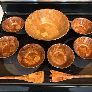 Pu&ograve; includere: Un set di sette ciotole di legno e un utensile da portata in legno. Le ciotole sono intrecciate e hanno una finitura in legno naturale. L'utensile da portata ha un manico lungo e una testa piatta e rettangolare.