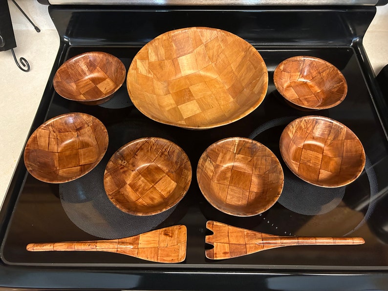 Pu&ograve; includere: Un set di sette ciotole di legno e un utensile da portata in legno. Le ciotole sono intrecciate e hanno una finitura in legno naturale. L'utensile da portata ha un manico lungo e una testa piatta e rettangolare.