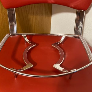 May include: A chrome-plated metal bar stool footrest with a spring-loaded design. The footrest is attached to the base of the stool and has a curved shape.