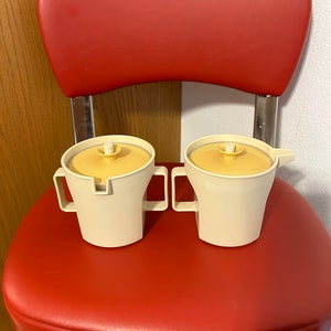 May include: Two white ceramic creamer and sugar containers with yellow lids. The containers are sitting on a red chair.
