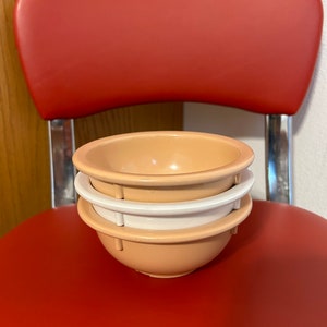 May include: Three nesting bowls in shades of peach and white. The bowls are stacked on a red chair.