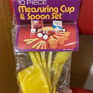 May include: A 10-piece measuring cup and spoon set in a clear plastic bag. The packaging is purple and features the text "10 PIECE Measuring Cup & Spoon Set" in yellow. The set includes yellow measuring cups and spoons.