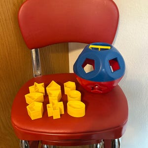 May include: A blue and red spherical shape-sorting toy with a yellow handle, accompanied by a collection of yellow plastic geometric blocks including a star, triangle, hexagon, square, cross, and oval, designed for matching into the sphere.