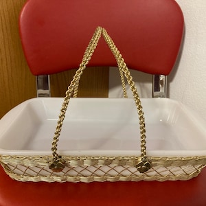 May include: White ceramic baking dish with a gold wire handle and gold accents. The dish is sitting on a red chair.