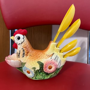 May include: A ceramic hen-shaped utensil holder with yellow spoons. The hen is painted with orange, yellow, and brown tones, featuring a red comb and floral accents. The holder is designed to hold the included yellow spoons.