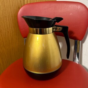 May include: A vintage gold and black insulated coffee carafe with a black handle and lid. The carafe has a gold band around the top and sits on a red chair. The carafe is designed to keep coffee hot.