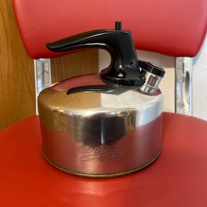 May include: A vintage, stainless steel kettle with a black handle and spout. The kettle has a rounded body and a small, silver-colored cap. The words "Cook's Romance" are etched on the side. The kettle is sitting on a red surface.