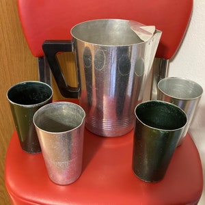 May include: A set of four aluminum cups and a large aluminum pitcher with a black handle. The cups and pitcher have a silver finish and are sitting on a red surface.