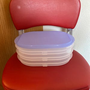 May include: A stack of four rectangular, clear plastic food storage containers with rounded corners. Each container features a translucent, light purple snap-on lid. The containers are stacked vertically, creating a uniform, layered appearance.