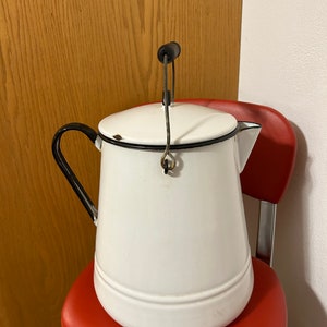 May include: White enamelware pitcher with a black rim and handle. The pitcher has a wire bail handle with a wooden knob. The pitcher is sitting on a red chair.