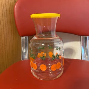 May include: A clear glass pitcher with a yellow lid and orange and green fruit design.